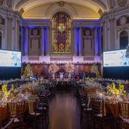 Elegant banquet hall with decorated tables, stained glass window, and large screens for an event.