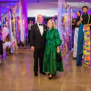 Couple posing at event with performers on stilts holding colorful decorations in brightly lit space.