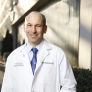 A smiling doctor in a lab coat stands outside a building, wearing a colorful tie.