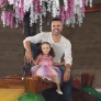 A smiling man and a young girl in colorful clothing pose together on a wooden bench under floral decorations.