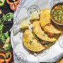Three tacos with lime wedges and a small bowl of salsa, surrounded by fresh herbs and vegetables.