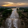 Aerial view of a river with flowing water at sunset, surrounded by greenery and a small settlement.