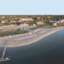 Aerial view of a beachfront with lounge chairs, cabanas, a pier, and houses in the background.
