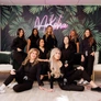 Group of eight women posing indoors in front of a leafy wall with a neon Akasha sign.