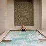 Woman relaxing in an indoor pool with tiled wall and beige stone surroundings.