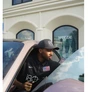 Man wearing a black cap and shirt sitting in a car, cleaning the windshield outside a building.