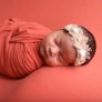 Newborn baby peacefully sleeping, wrapped in orange cloth with a decorative headband, on a matching orange background.