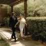 Bride in white dress and groom in black suit walking hand in hand outdoors under wooden pergola.