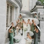 Bride with bridesmaids in green dresses holding white parasols on stone staircase outside building.