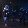 Band performing live on stage with female and male vocalists and instrumentalists in dim lighting.
