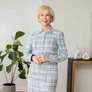 Portrait of a confident older woman in a light blue plaid suit standing indoors near plants.