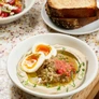 Bowl of hummus topped with soft-boiled egg, tomato, herbs, and a side of toasted bread and salad.