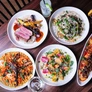 Table with five diverse gourmet dishes and two drinks, viewed from above in a restaurant setting.