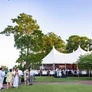 Outdoor event with people socializing near a large white tent by the water and trees.