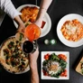 Top view of a table with Italian dishes including pizza, pasta, salad, and drinks being toasted by hands.