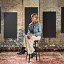 Woman sitting on a stool in a rustic room with brick walls, black panels, and a patterned carpet.