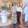 Two smiling women standing in a boutique clothing store with displayed clothes and decor.