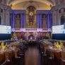 Elegant banquet hall with decorated tables, stained glass window, and large screens for an event.