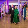 Couple posing at event with performers on stilts holding colorful decorations in brightly lit space.