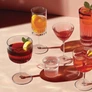 Various colorful cocktails and a glass of water on a light pink surface with soft shadows.