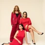 Three women in red outfits posing against beige background, one standing, one seated, one reclining.