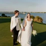 Bride in white gown and groom in white tuxedo walking hand in hand on grass near water.