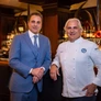 Two men, one in a blue suit and the other in a white chef's jacket, standing in a bar setting.