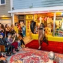 Drag performer walking on outdoor runway with seated and standing audience in front of colorful storefront.