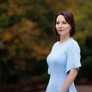 Woman in light blue dress standing outdoors with blurred trees in the background.
