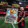 Man in Atlanta United jersey holding colorful Atlanta-themed artwork inside Mercedes-Benz Stadium.