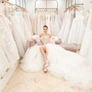 Woman in an elaborate white wedding dress sitting in a bridal shop surrounded by wedding gowns.