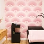 Room with pink eye-patterned wallpaper, black chair with pink blanket, wooden table, and black lamp.