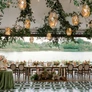 Elegant outdoor dining setup with hanging lanterns, greenery, floral centerpieces, and a lakeside view.