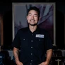Smiling man with tattoo wearing a black shirt with ADALINA name tag in a dimly lit restaurant.