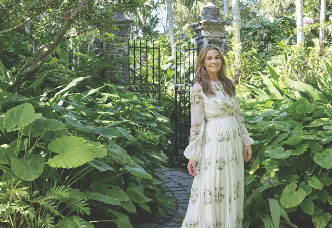 A woman in a floral dress stands amidst lush greenery near a garden gate.
