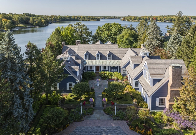 Aerial view of a large blue house surrounded by greenery, overlooking a calm lake.