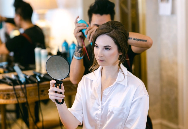 A woman in a white shirt mirrors her hairstyle while a stylist works behind her.