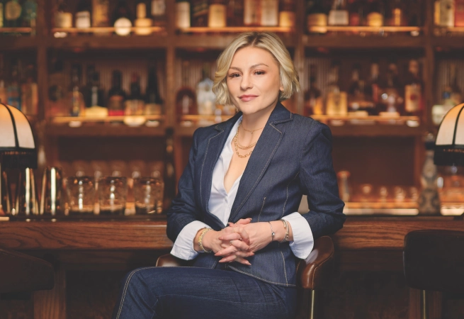 A woman with short blonde hair sits confidently in a bar, wearing a denim suit and looking directly at the camera.