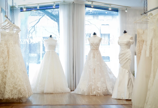Display of elegant wedding dresses on mannequins inside a bridal boutique, with large windows and white curtains.