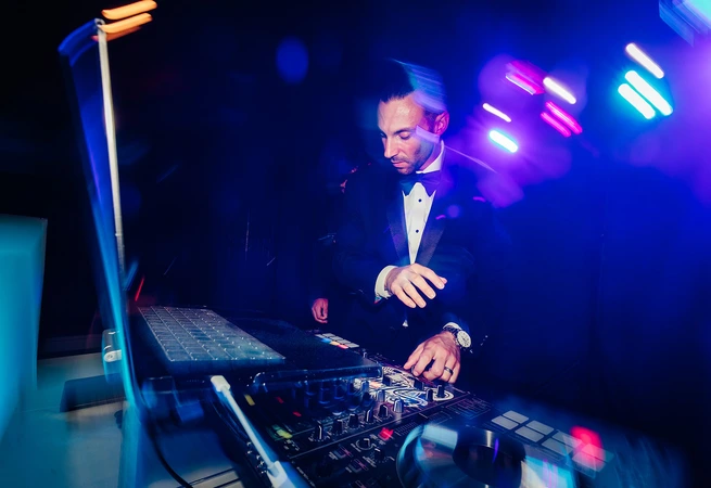 Man in tuxedo DJing at a nightclub with colorful lights and motion blur effects.