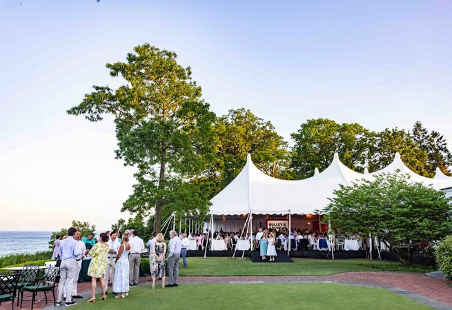 Outdoor event with people socializing near a large white tent by the water and trees.