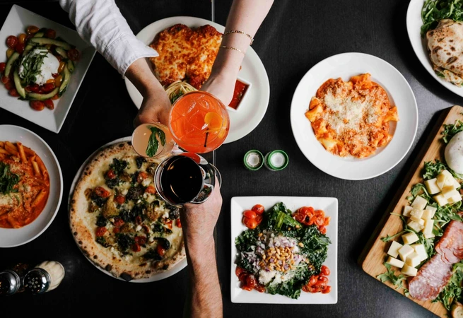 Top view of a table with Italian dishes including pizza, pasta, salad, and drinks being toasted by hands.