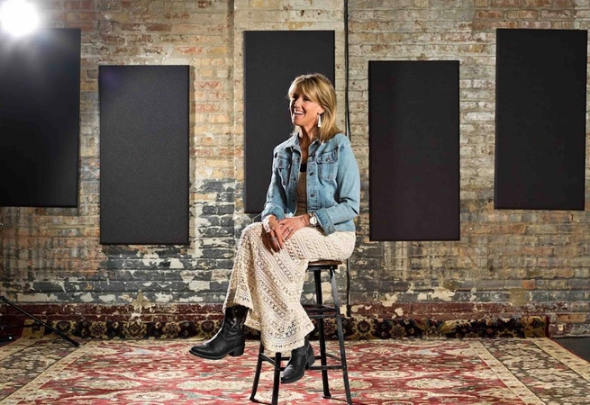 Woman sitting on a stool in a rustic room with brick walls, black panels, and a patterned carpet.