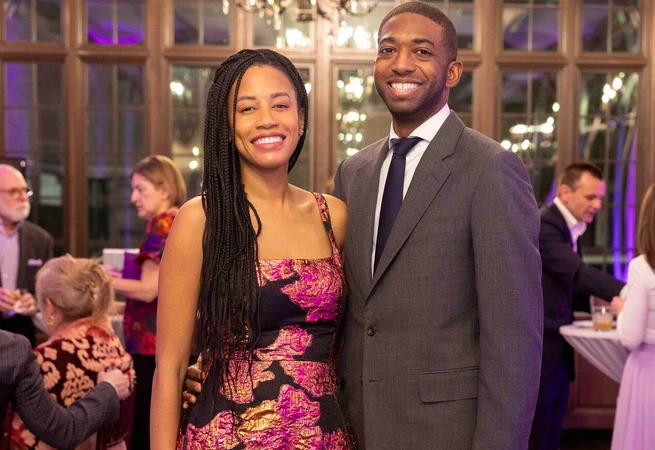 A smiling couple dressed formally at an indoor event with guests in the background.