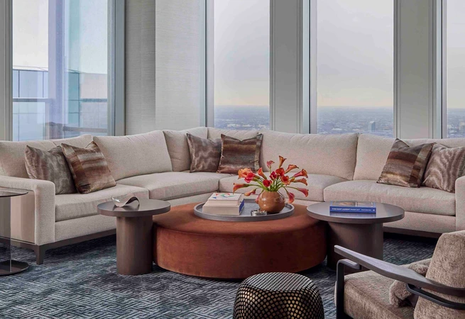 Modern living room with large beige sectional sofa, round coffee table with flowers, and panoramic city view windows.