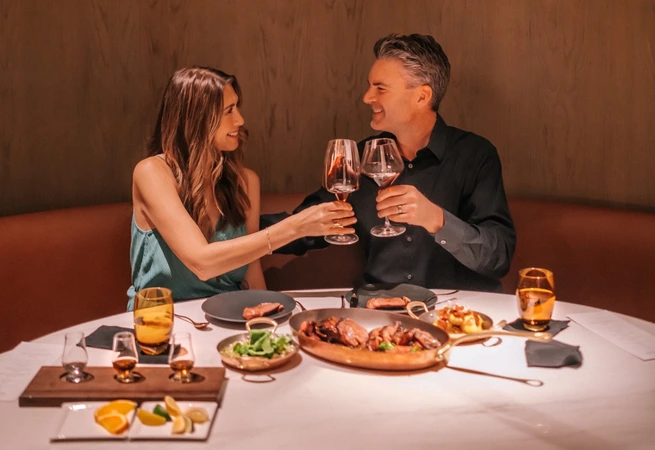 Two people clinking wine glasses over a dinner table with food in a restaurant.