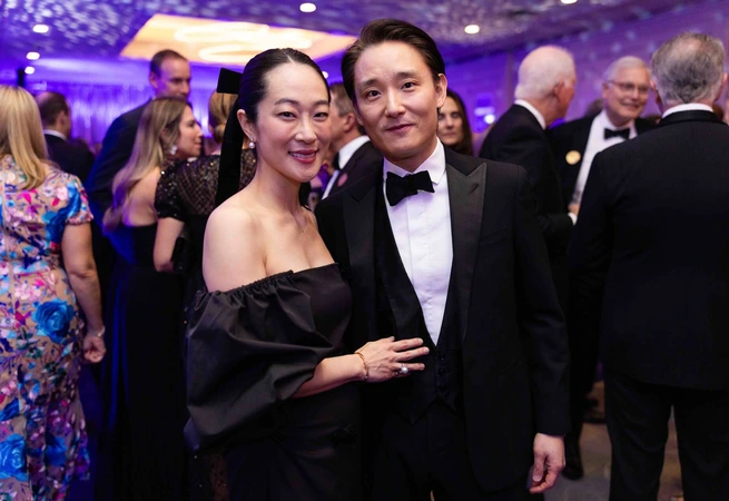 Couple dressed formally, man in tuxedo, woman in black off-shoulder dress, at elegant indoor event with guests.