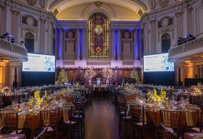 Elegant banquet hall with decorated tables, stained glass window, and large screens for an event.