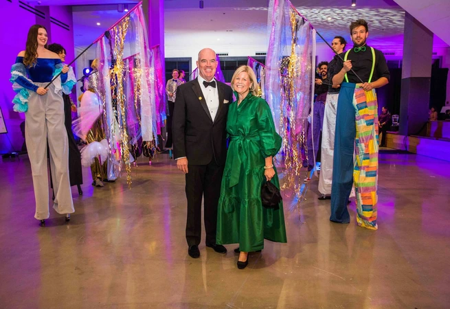 Couple posing at event with performers on stilts holding colorful decorations in brightly lit space.