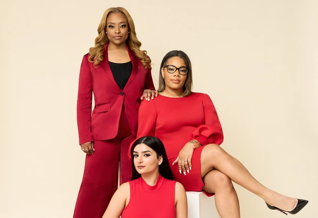 Three women in red outfits posing against beige background, one standing, one seated, one reclining.